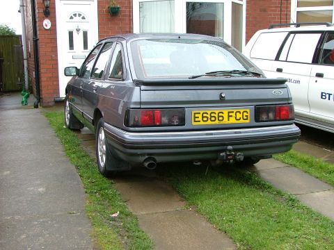 1988 Ford Sport - The Ford Sierra hatchback is parked on a driveway in front of a house, with a white van to its right.