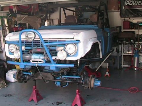 1971 Ford Bronco - A blue and white Ford Bronco is undergoing repairs in a garage, elevated on red jack stands.