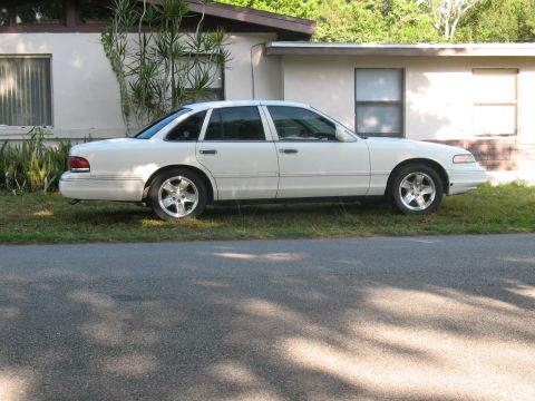 1997 Ford Crown Victoria - A white Ford Crown Victoria full-size sedan shown in profile from the driver's side, parked on grass in front of a single-story house. It h…