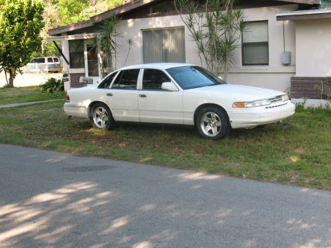 1997 Ford Crown Victoria - A white Ford Crown Victoria four-door sedan parked on a grassy lawn in front of a single-story stucco house, shown from a front three-quart…