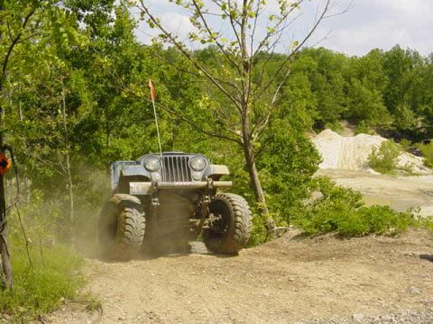 1992 Ford Bronco - Lifted flat-fender Willys-style Jeep with a flag on large off-road tires climbing a wooded dirt trail.