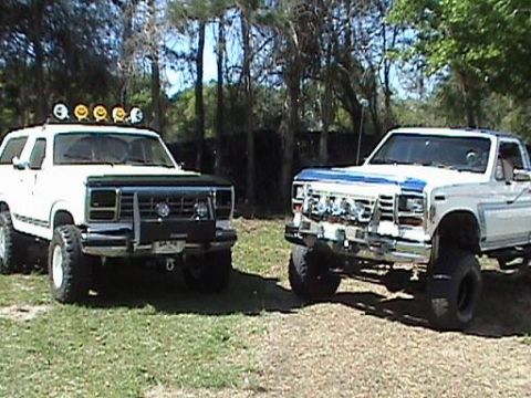 1986 Ford Bronco - Two lifted trucks, a white SUV and a white pickup, are parked outdoors on a sunny day.