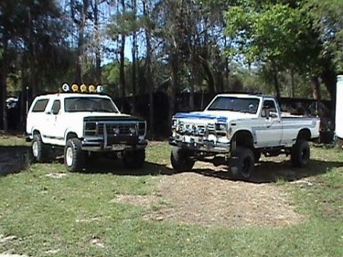 1986 Ford Bronco - Two lifted pickup trucks are parked outdoors in a grassy area with trees in the background.