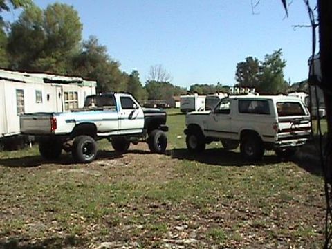 1986 Ford Bronco - A white 1986 Ford Bronco and a lifted pickup truck are parked in a grassy area with trailers in the background.