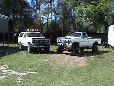 1986 Ford Bronco - Two lifted trucks are parked side-by-side in a yard with trees in the background.