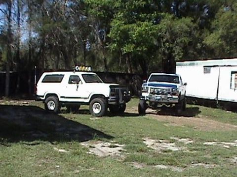 1986 Ford Bronco - A white 1986 Ford Bronco with auxiliary lights on the roof sits in a grassy area next to a lifted pickup truck.