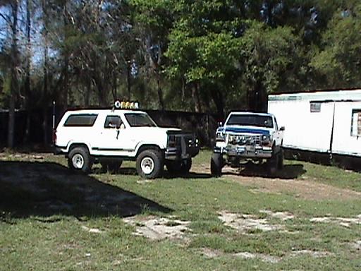White 1986 Ford Bronco with off-road lights, parked next to a lifted pickup truck