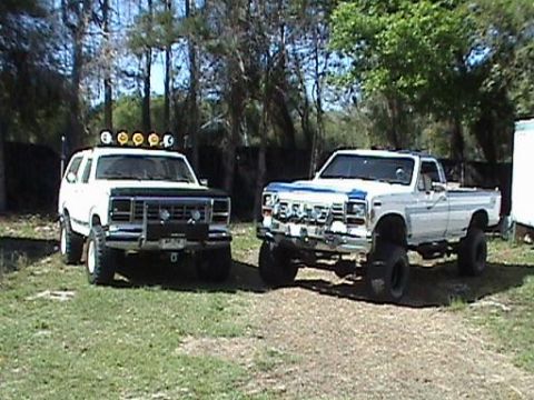 1986 Ford Bronco - Two lifted 1980s Ford trucks, a Bronco and a pickup, are parked outside on a sunny day.