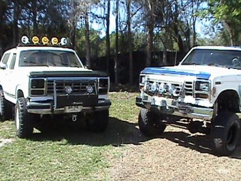 1986 Ford Bronco - Two modified Ford trucks are parked side-by-side on a grassy area with trees in the background.