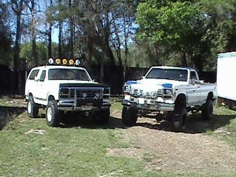 1986 Ford Bronco - Two modified trucks, a white SUV and a white pickup, are parked outdoors on a sunny day.