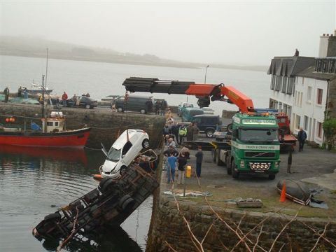 1989 Ford Bronco - A crane truck is actively recovering a white car that has fallen into the water near a harbor.