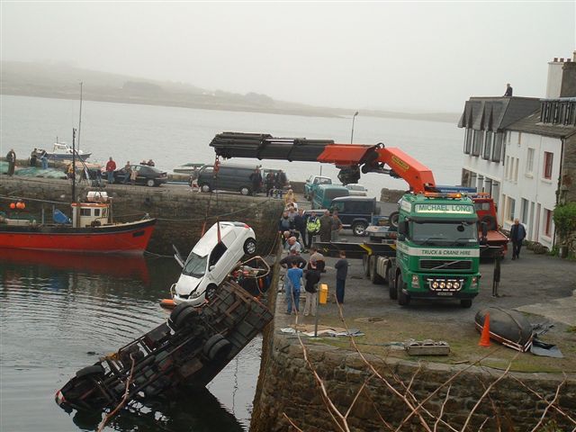 Crane truck lifting a white car from water next to a harbor wall