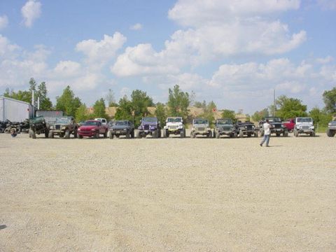 1992 Ford Bronco - A group of off-road vehicles are lined up in a gravel lot under a partly cloudy sky.