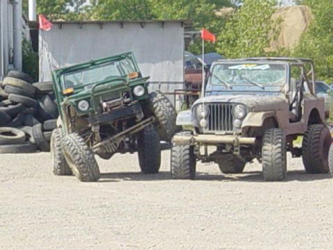 1992 Ford Bronco - Two off-road vehicles are parked side-by-side on a gravel surface with a building in the background.