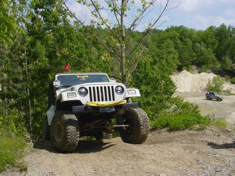 1992 Ford Bronco - white lifted Jeep Wrangler on oversized mud tires with a yellow tow strap sits on a wooded gravel trail.