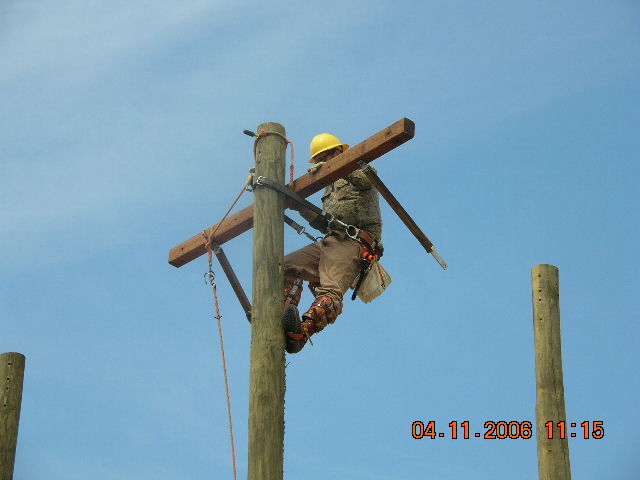 Lineman in yellow hard hat climbing a wooden utility pole with crossbars, blue sky