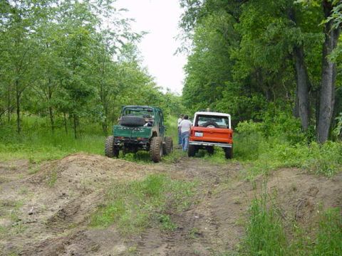 1992 Ford Bronco - Two off-road vehicles are parked on a dirt path surrounded by lush green trees.