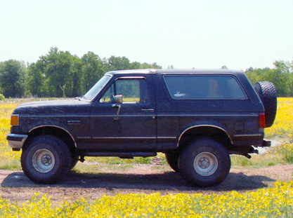 1988 Ford Bronco - A dark blue 1988 Ford Bronco is seen from the side, parked in a grassy field with trees in the background.