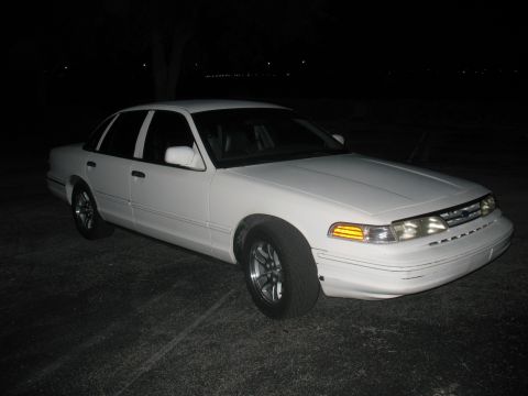 1997 Ford Crown Victoria - A white 1997 Ford Crown Victoria is parked outside at night with its headlights on.
