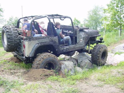 1992 Ford Bronco - An off-road vehicle with a roll cage and large tires is shown traversing a rocky and muddy terrain.