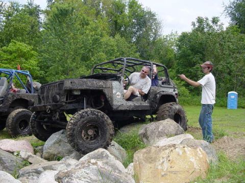1992 Ford Bronco - Two off-road vehicles are parked on rocks with trees in the background and a portable toilet to the right.