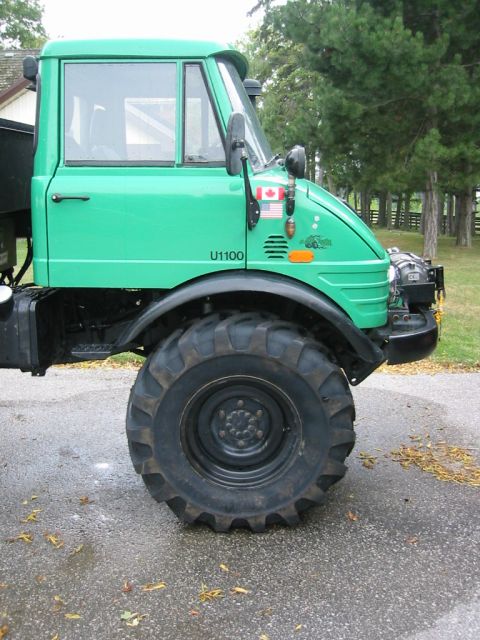1991 Ford Bronco - A green Unimog U1100 truck is parked on a wet asphalt surface with trees in the background.