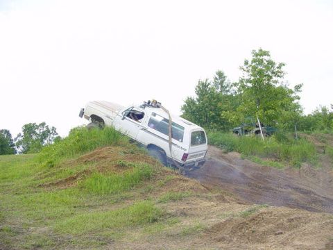 1992 Ford Bronco - A white SUV is attempting to ascend a muddy, grassy incline, with trees visible in the distance.