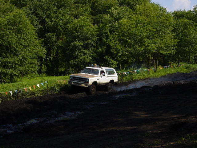 White SUV with brown hood driving through mud, trees in background