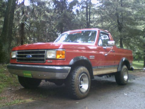 1989 Ford Bronco - This red Ford Bronco is parked off-road, surrounded by trees. It features a two-tone paint scheme.