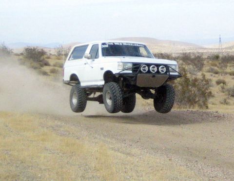 1990 Ford Bronco - white lifted Ford Bronco with off-road lights and a tube bumper jumps and gets airborne on a dirt road in the desert.