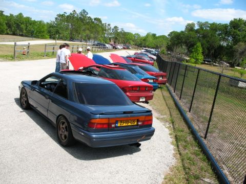 2006 Mazda 6 - A line of cars are parked next to a chain-link fence on a sunny day.