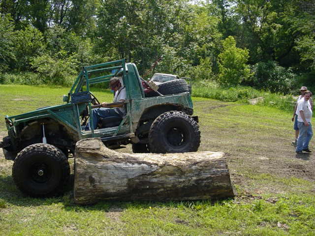 Green off-road vehicle with a roll cage and large tires, positioned near a fallen log