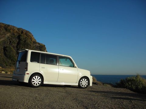 2006 Scion xB - A white 2006 Scion xB is parked on a gravelly cliff overlooking the blue ocean under a clear sky.