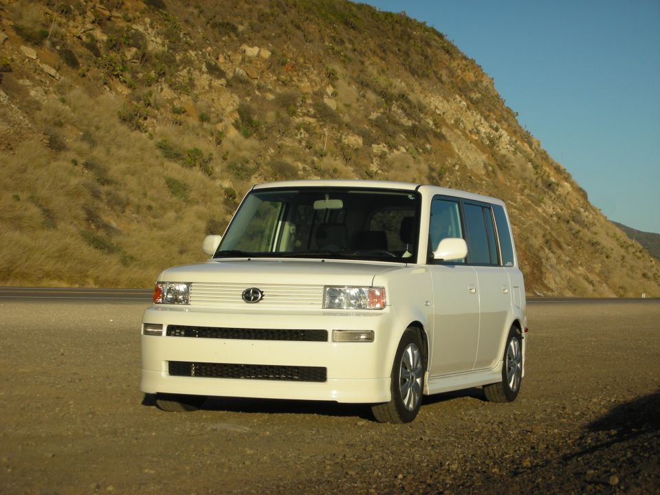 White Scion xB boxy wagon, front 3/4 view, parked on gravel near a hillside
