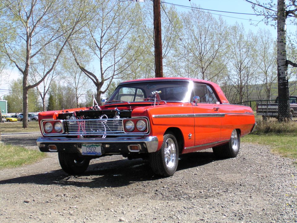 Red coupe with hood off showing chrome V8 engine, parked on gravel near trees