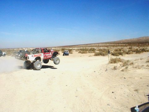 1996 Ford Bronco - A custom off-road truck soars through the air during a desert race, with other vehicles visible in the background.
