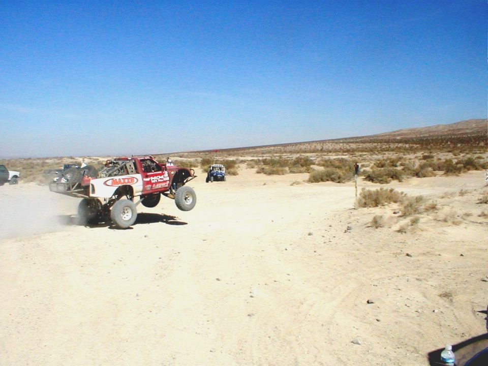 Red and white off-road truck mid-air jump, desert terrain, blue sky