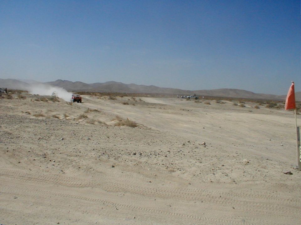 Red off-road vehicle kicking up dust on a dirt track in a desert with other vehicles in the distance