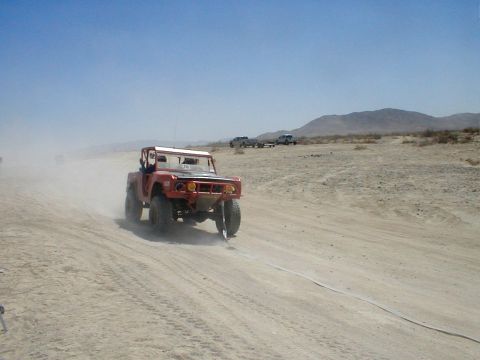 1996 Ford Bronco - A modified off-road vehicle drives through a dusty desert landscape, with other vehicles visible in the distance.