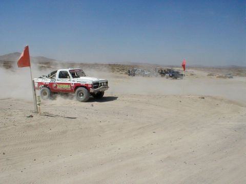 1996 Ford Bronco - A white and red off-road truck races across a dusty desert landscape with other vehicles in the background.