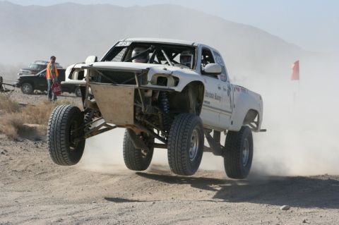 1996 Ford Bronco - Off-road truck mid-air during a race, kicking up dust on a desert course.