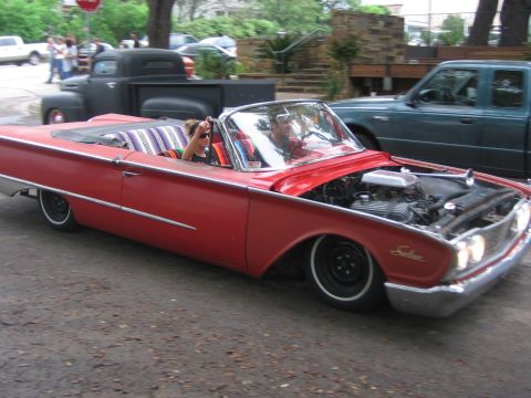 1996 Ford Bronco - A red convertible cruises down a street with its hood open, revealing the engine.