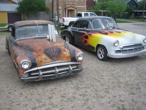 1996 Ford Bronco - Two vintage cars, one with a rusty patina and exposed exhaust pipes, the other with a vibrant flame paint job, parked side-by-side.