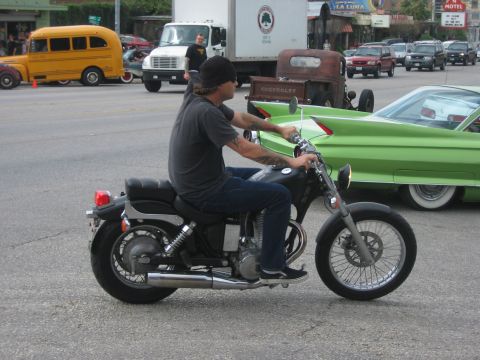 1996 Ford Bronco - A rider on a black motorcycle cruises past a bright green classic car on a street with other vehicles.