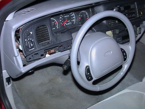 2000 Ford Crown Victoria - Driver's side of a gray Ford sedan interior with the dash bezel partly disassembled, the headlight switch panel and center vent hanging loo…