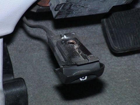 2000 Ford Crown Victoria - Close-up in the driver's footwell of the black parking-brake release handle, unbolted and hanging by its cable below the lower dash, with d…