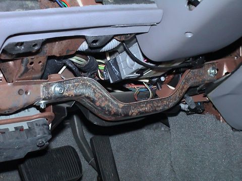 2000 Ford Crown Victoria - Under-dash close-up on the driver's side showing the surface-rusted knee bolster/steering column support bar bolted across the footwell, wi…