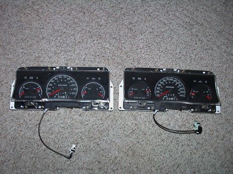 2000 Ford Crown Victoria - Two Ford instrument clusters laid side by side face-up on beige carpet for comparison: the one at left reads to 120 mph, the one at right t…