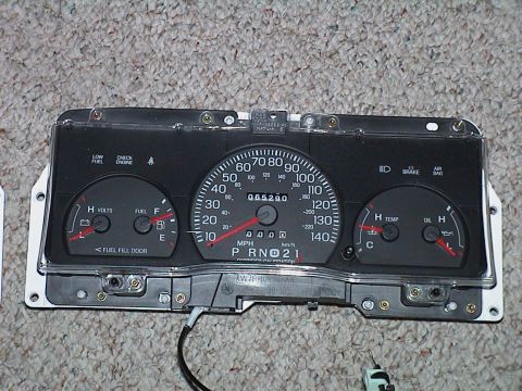 2000 Ford Crown Victoria - A 140 mph Ford instrument cluster lying face-up on carpet, with volts and fuel gauges at left, temp and oil pressure gauges at right, a 65,…