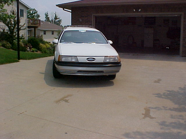 White first-generation Ford Taurus sedan photographed head-on in a driveway by an open garage